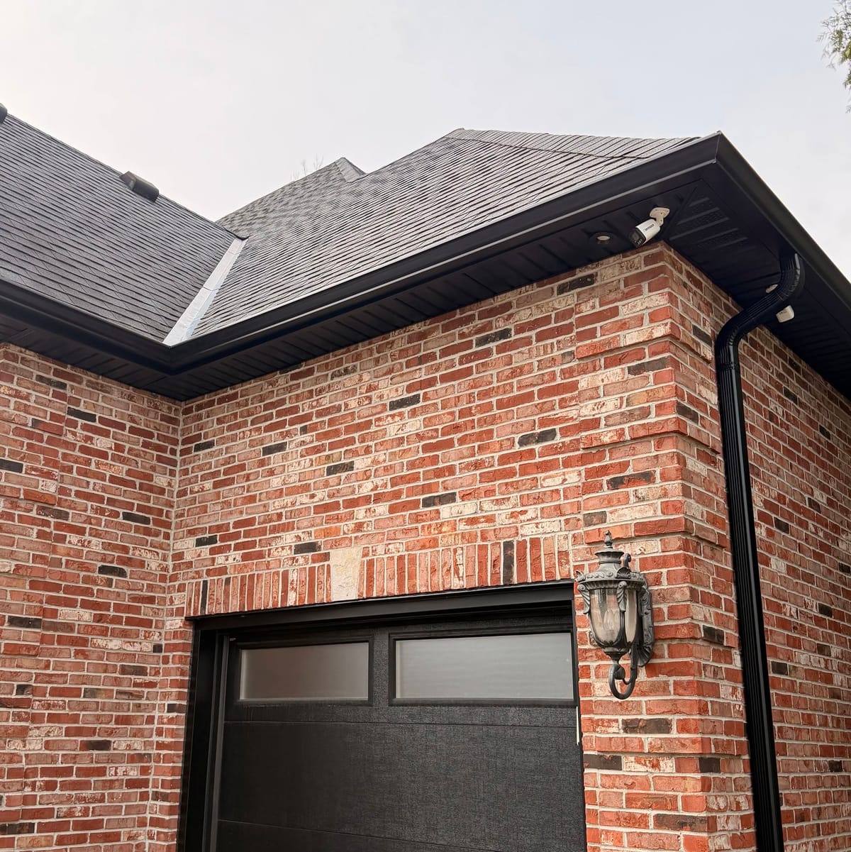Black soffit, fascia and seamless eavestrough along the roof edge of a red brick home, with a black downspout and garage door.