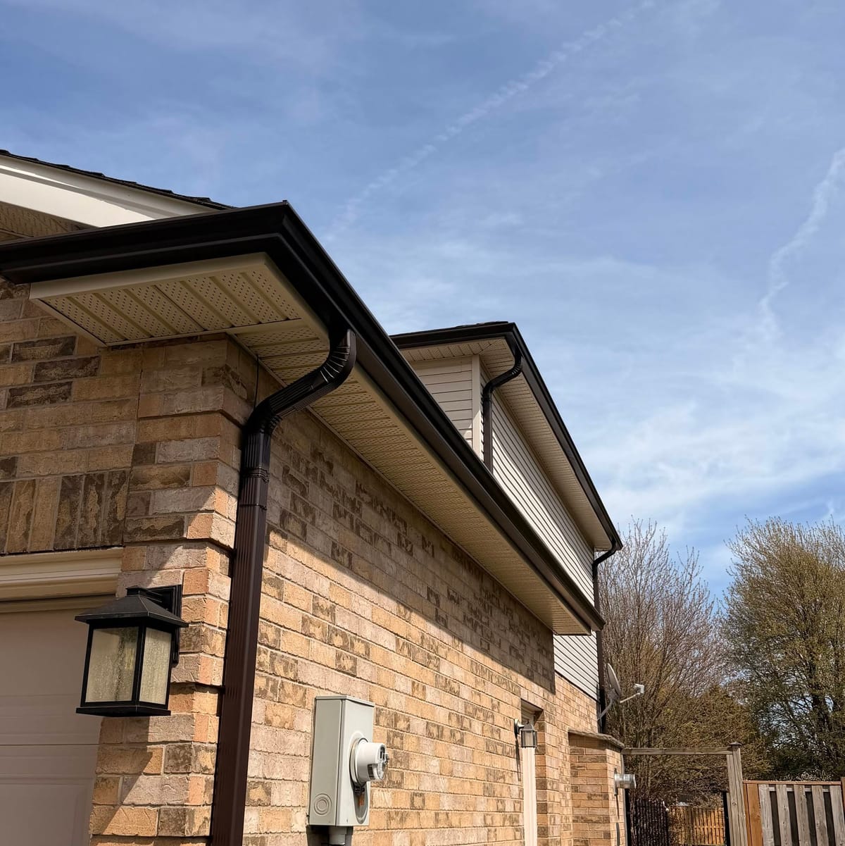 Dark brown seamless eavestrough and downspout on a tan brick home with beige siding above in the London, Ontario area