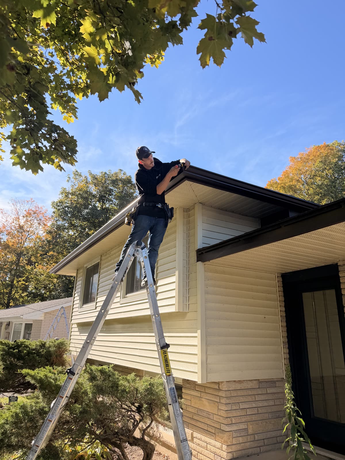 Forest City Eavestrough installer on a ladder fastening eavestrough to a house with cream siding