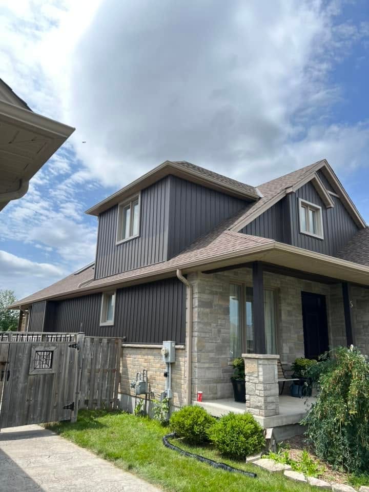 A storey-and-a-half home with dormers in dark grey vertical siding, with eavestrough and soffit, above a stone and brick base